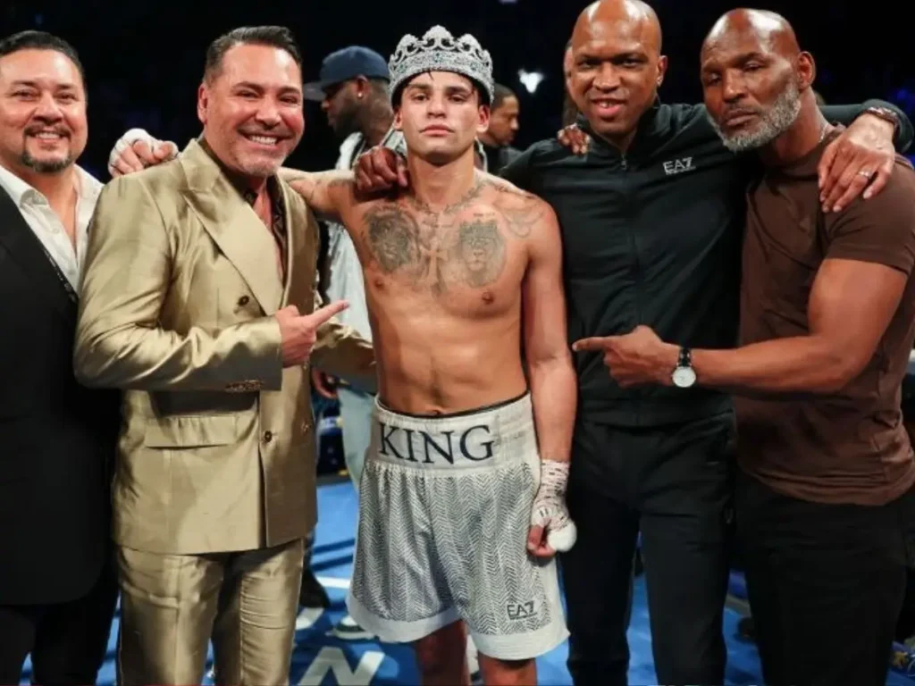 Ryan Garcia celebra vitória no ringue usando coroa e calção "King", ladeado pelo promotor Oscar De La Hoya e sua equipe. A imagem ilustra o momento de triunfo que deu início ao prazo de 90 dias imposto pelo WBC para a definição da próxima defesa de cinturão e as negociações de revanche